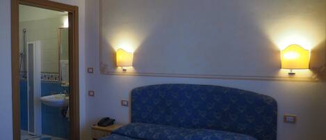 Una habitación de hotel con cama, mesitas de noche, un espejo y un baño visible a través de una puerta abierta.