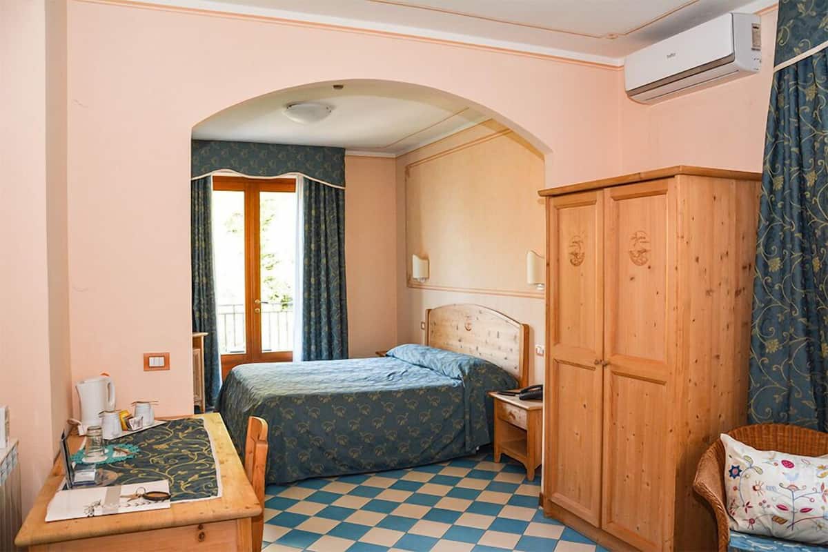 A bedroom with a checkered floor, a bed with a patterned bedspread, a wooden wardrobe, a desk with a chair, and a window with curtains.