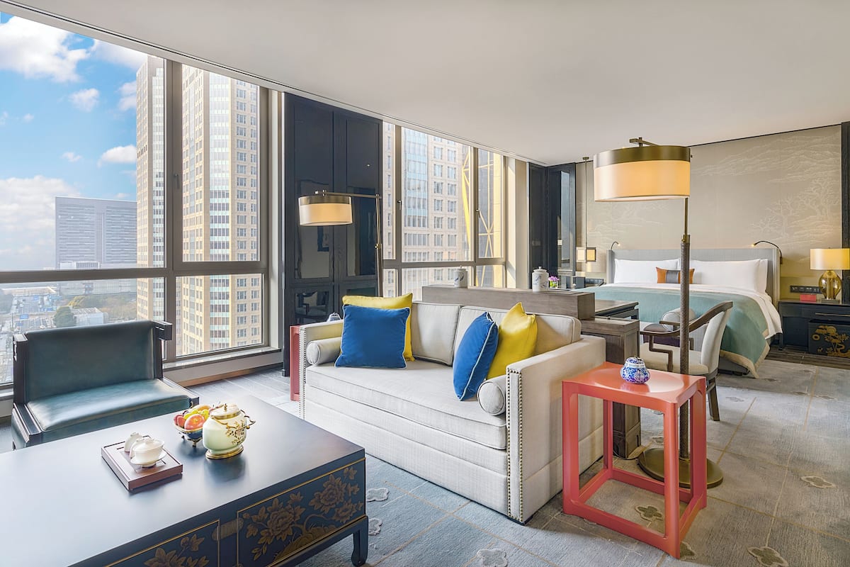 A modern hotel room with a large window, a bed, a sofa, a desk, and a nightstand.