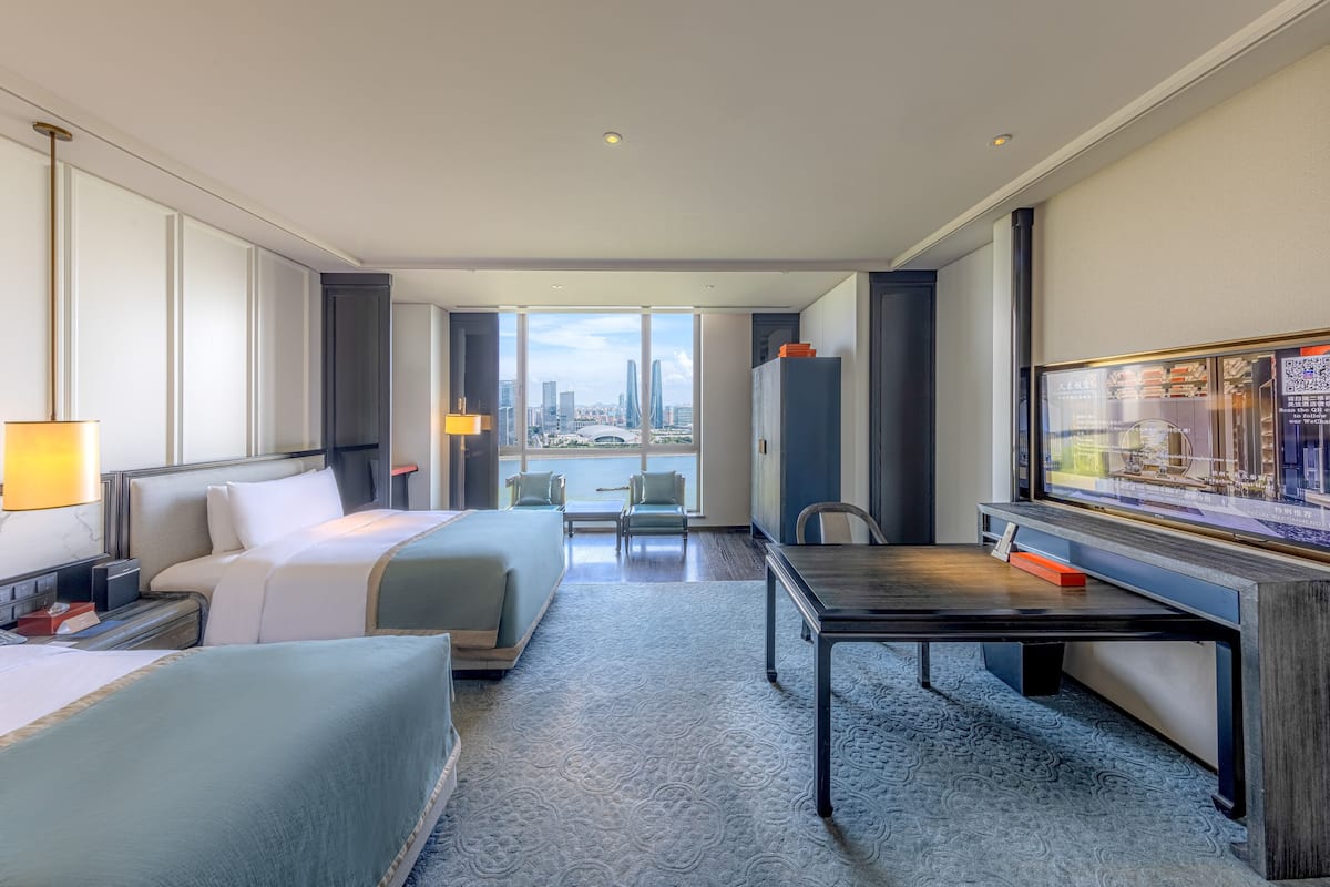 A hotel room with two beds, a desk, a TV, and a view of the city.