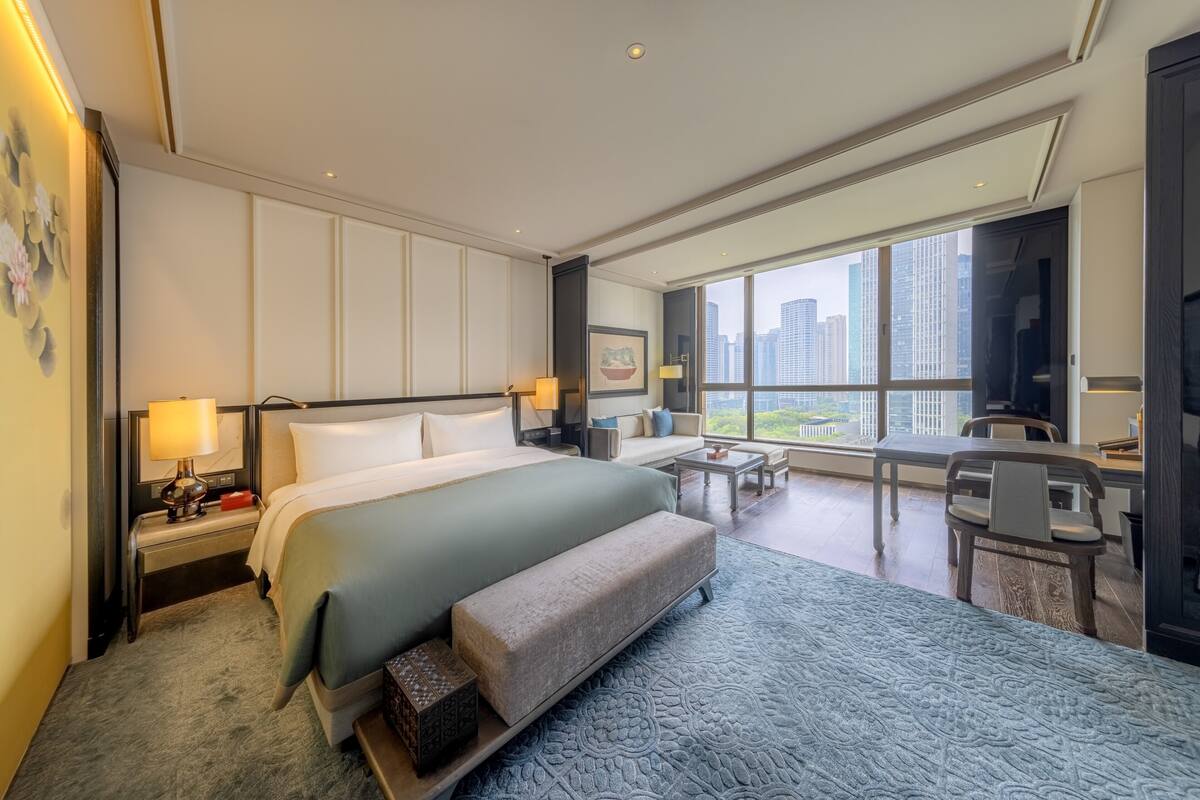 A modern hotel room with a large bed, a desk, a sofa, and a view of the city skyline.