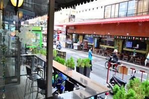 A street view from a window overlooking a restaurant with outdoor seating, a red awning, and a scooter parked outside. - Centro at Sansabai (Patong)