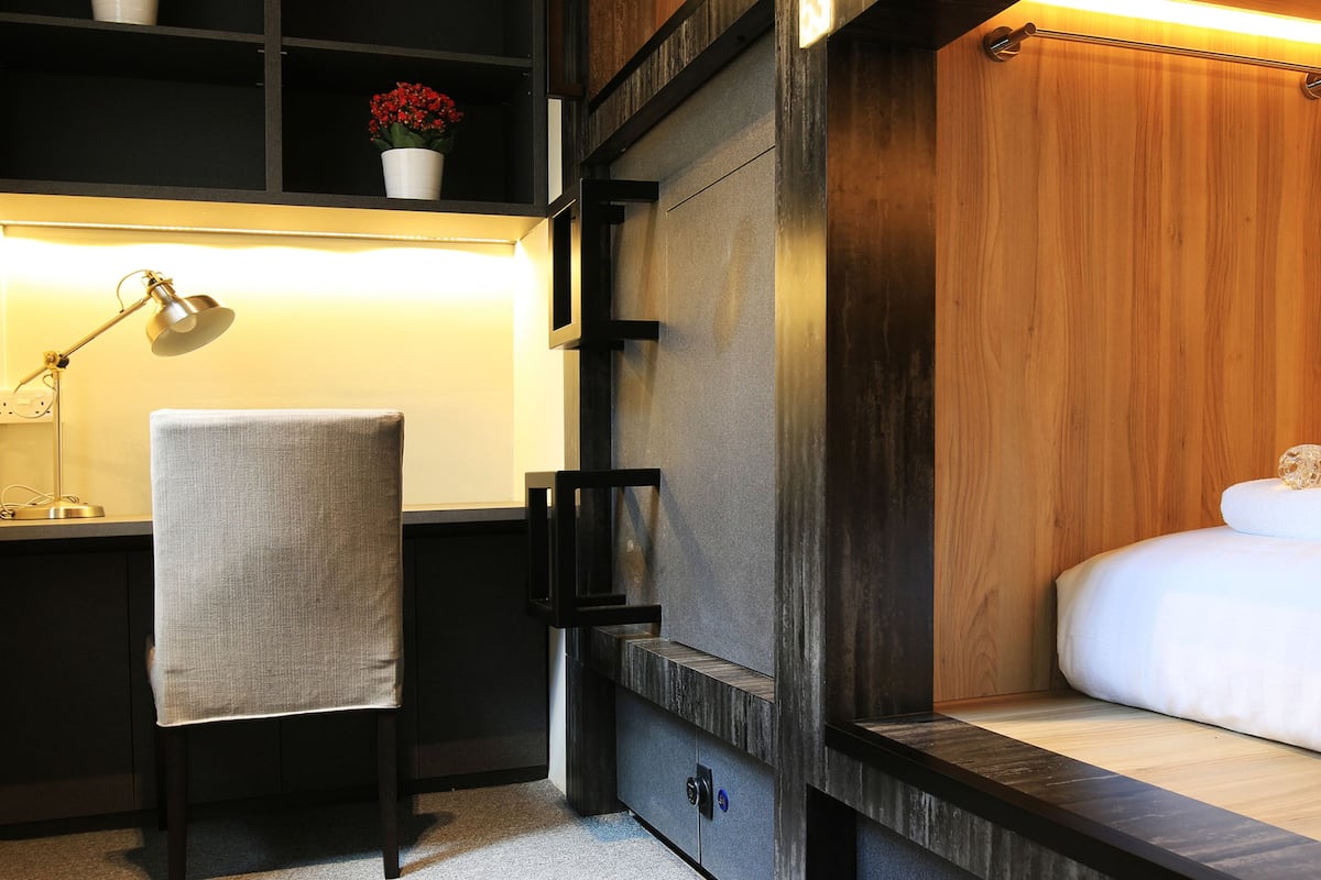 Quarto de hotel compacto com uma cama, uma escrivaninha com luminária, uma cadeira e uma estante com uma flor.
