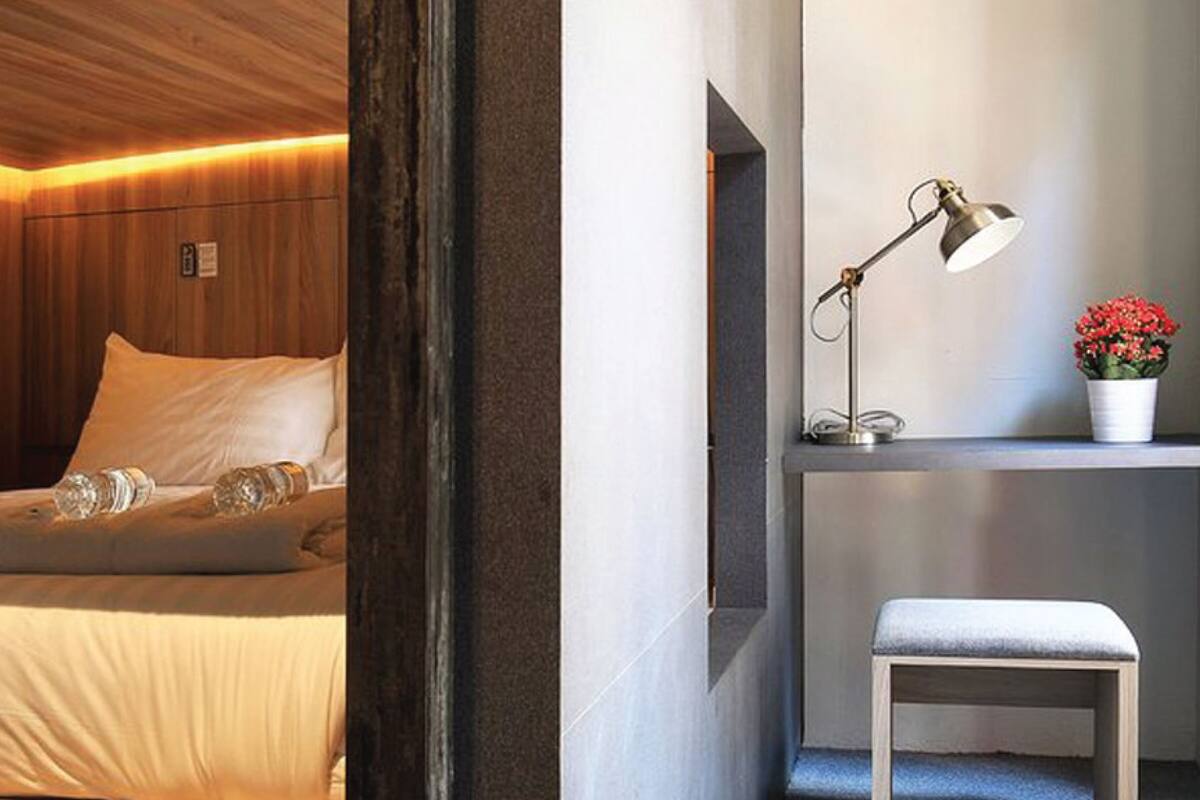 Quarto de hotel com cama, escrivaninha, luminária e um pequeno banco.