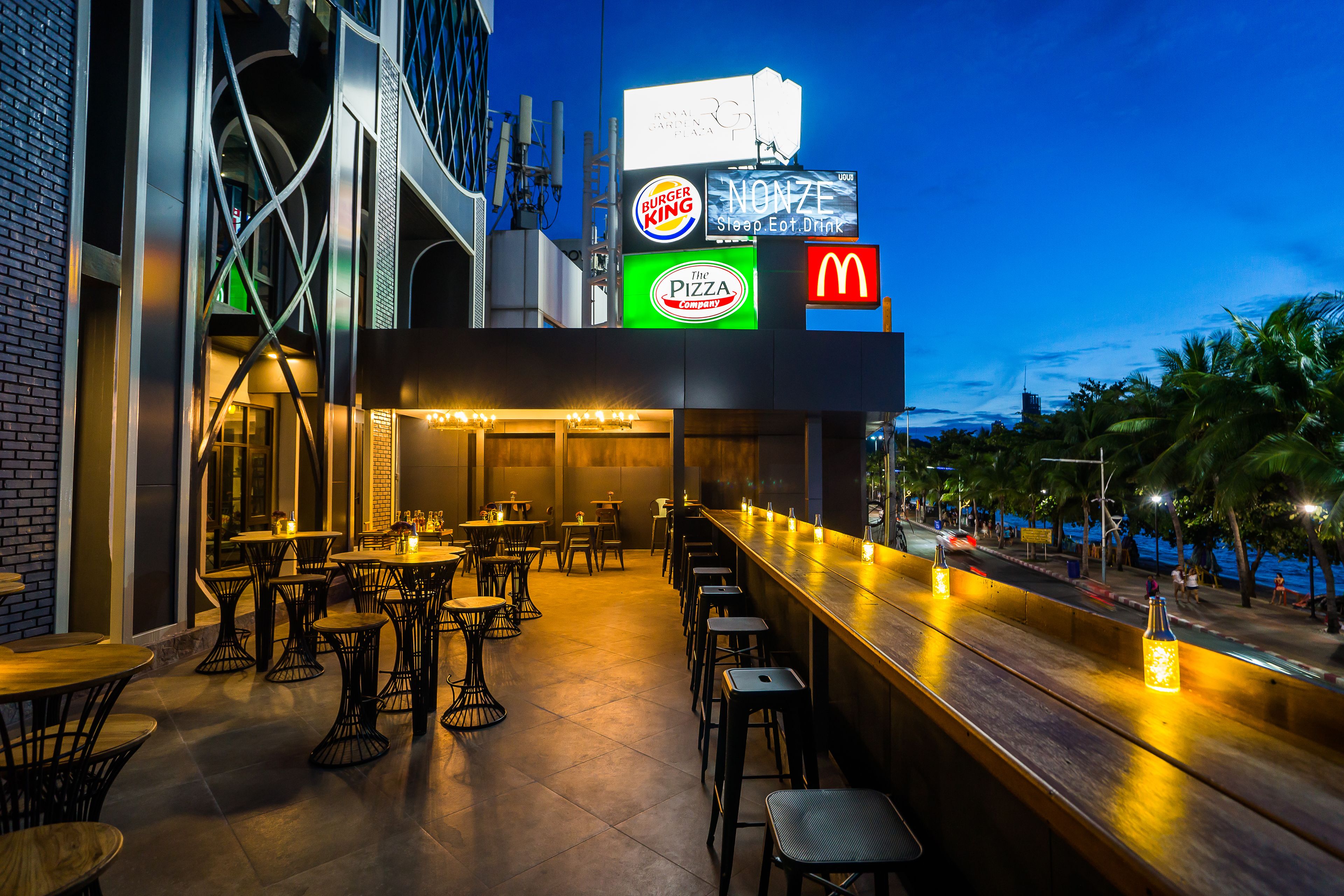 Zona de bar al aire libre con mesas y taburetes, iluminada con luces colgantes.