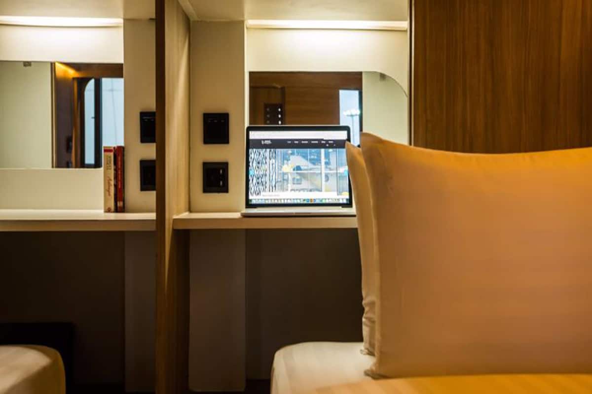 A hotel room with a bed, a laptop on a desk, and a window with a view of buildings.