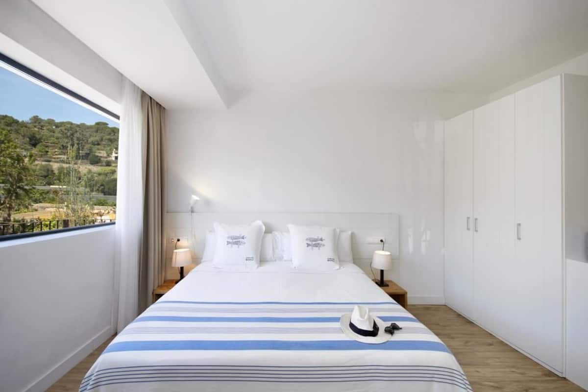 A modern hotel room with a large bed, a view of the outdoors, and white furniture.
