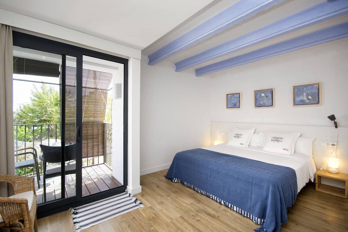 A bedroom with a large bed, a balcony with wicker furniture, and three framed artworks on the wall.