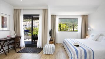 Une chambre d’hôtel comprenant un lit, un bureau, une chaise, une petite table, un téléviseur et un balcon donnant sur un paysage verdoyant et des bâtiments.
