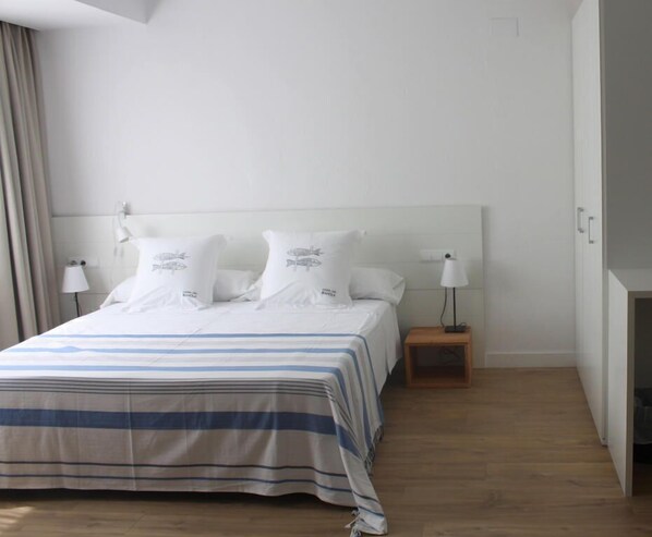 Un dormitorio con cama, almohadas blancas, una mesita de noche con lámpara y suelo de madera.