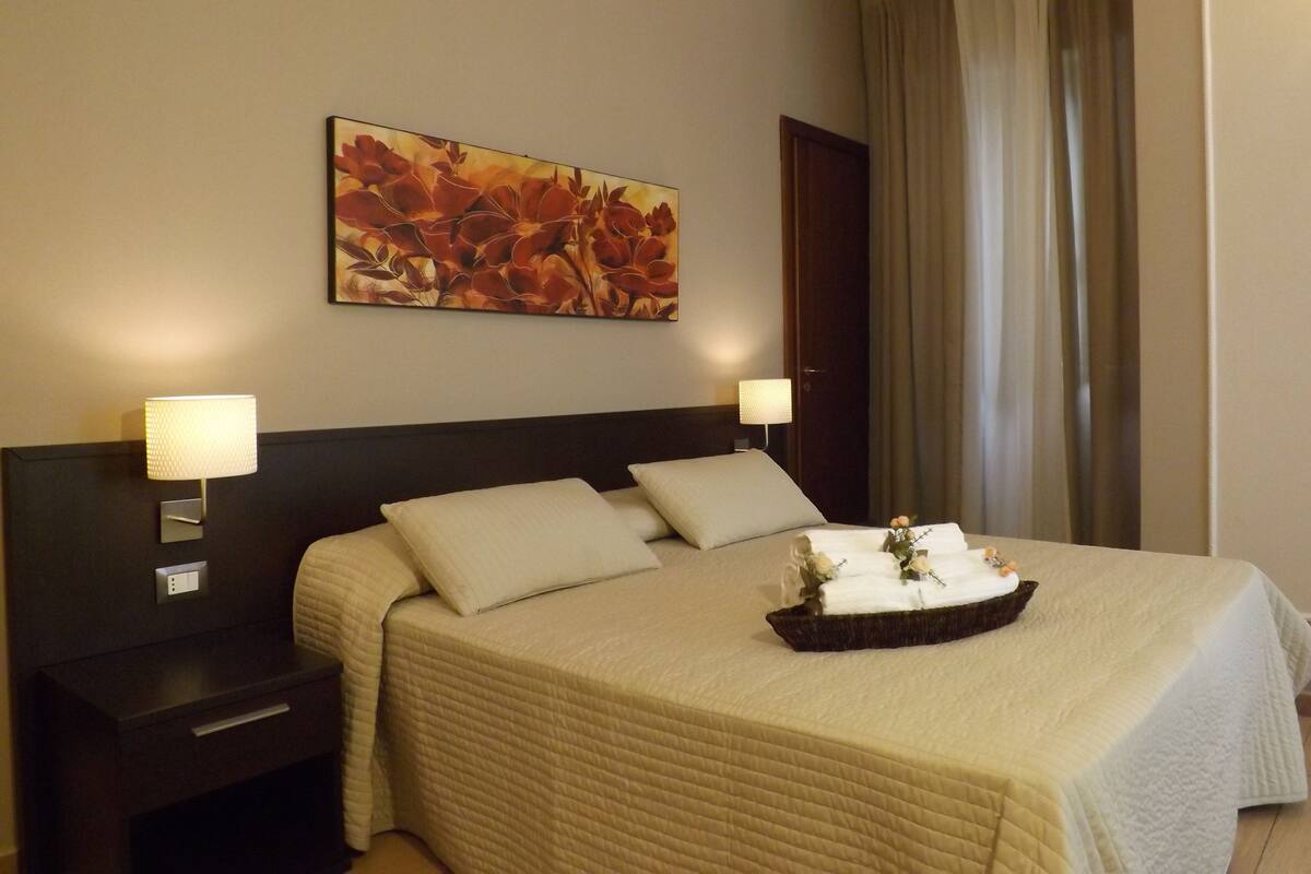 A hotel room with a bed, two bedside lamps, a nightstand, a wall-mounted painting, and a window with curtains.