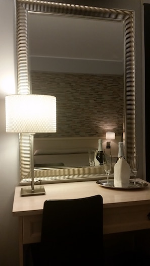 Habitación de hotel con un espejo grande, una mesita de noche con lámpara, una bandeja con botella y vasos, y una silla.