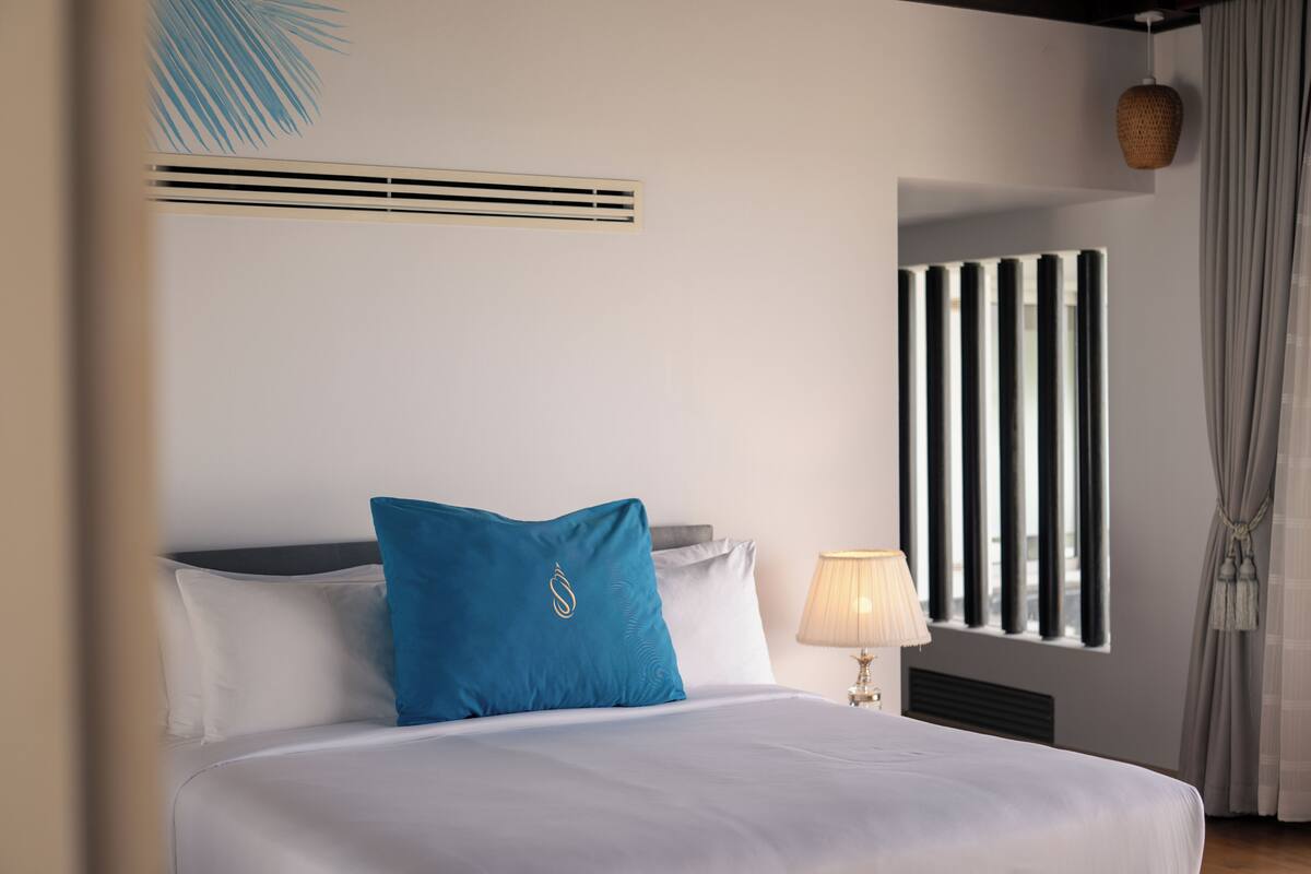 A hotel room with a bed, a window with curtains, a wall-mounted radiator, and a lamp.