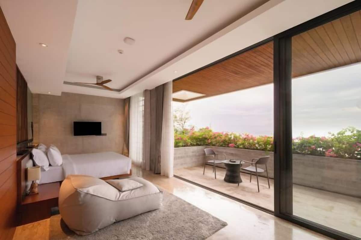 A modern bedroom with a bed, a flat-screen TV, a sofa, and a balcony with outdoor seating.