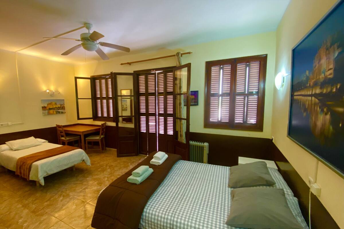 A hotel room with two beds, a desk, a ceiling fan, and a large window with shutters.