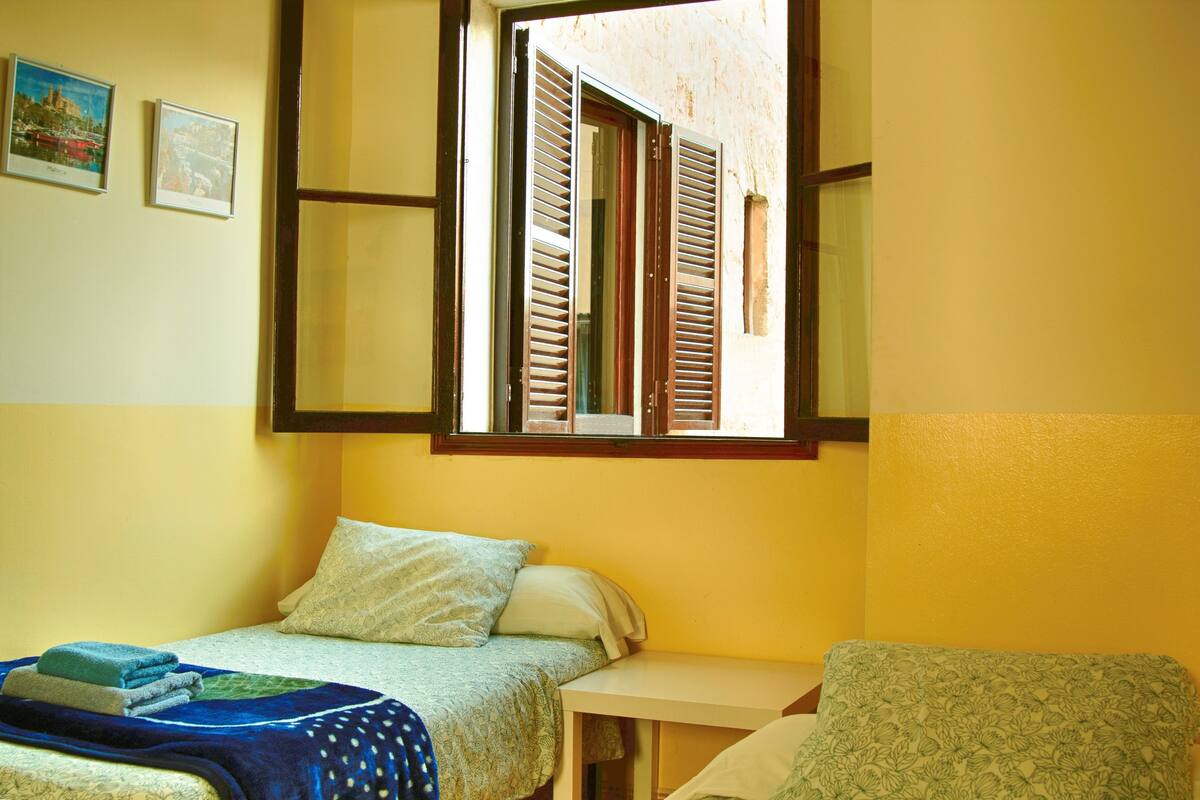 A small, bright room with a bed, a bedside table, and a window with shutters.