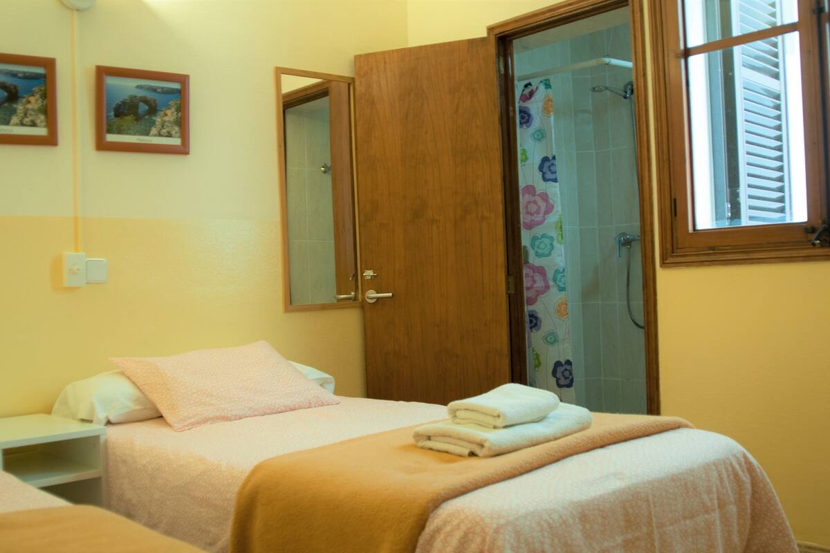 A hotel room with two beds, a wooden door, a shower with floral curtains, and a window with blinds.