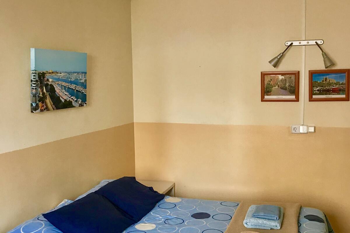 A bedroom with a bed, pillows, a blanket, a desk, and a chair. There are two framed pictures on the wall.
