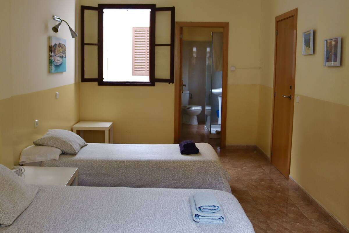A hotel room with two beds, a small table, a window with shutters, and a bathroom visible through an open door.