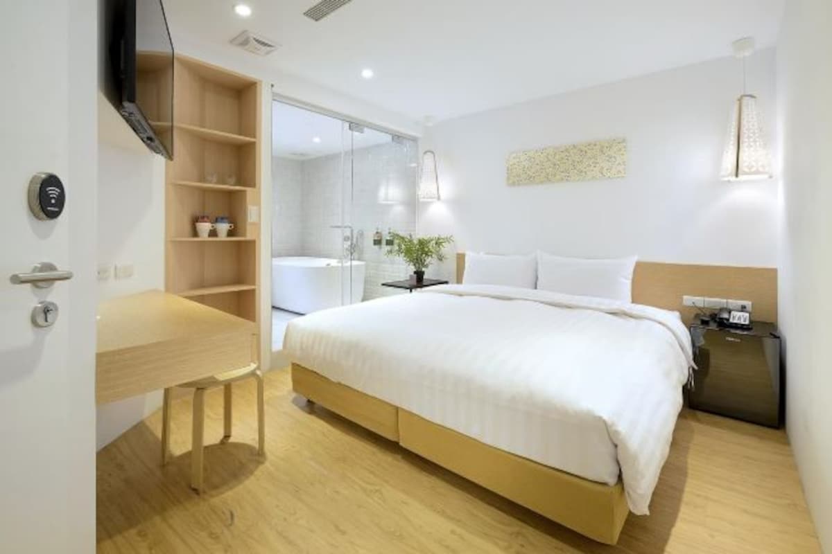 A modern hotel room with a large bed, a wooden desk, a built-in TV, a bedside table, and a mirror.