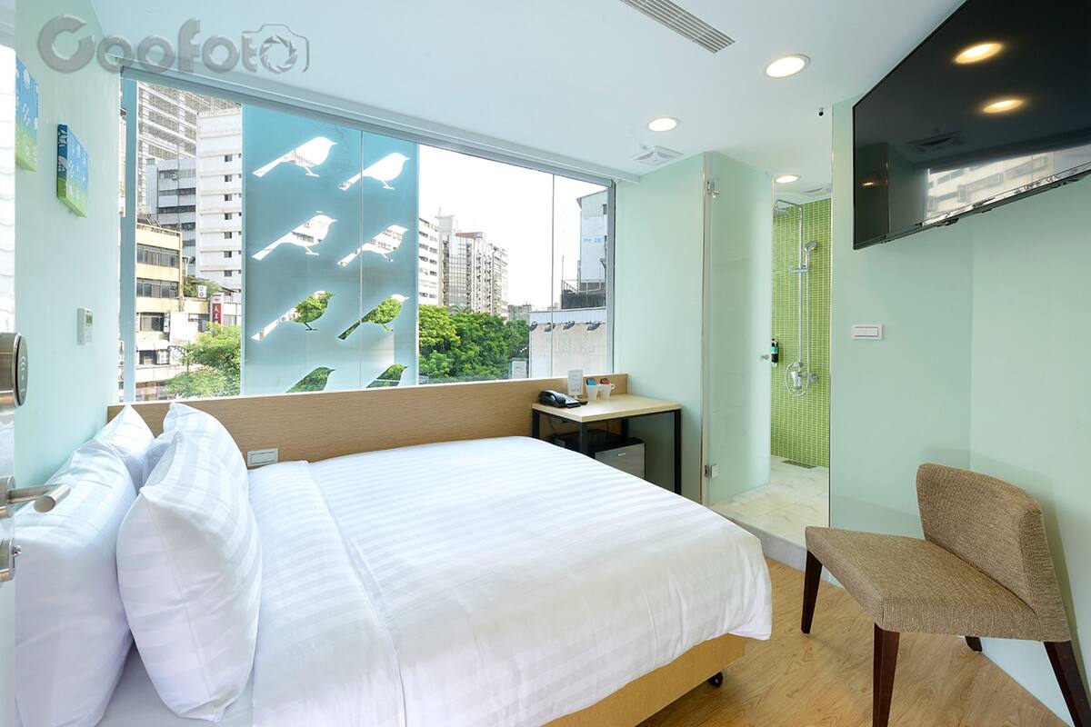 A hotel room with a large bed, a desk, a chair, a TV, and a window with a city view.