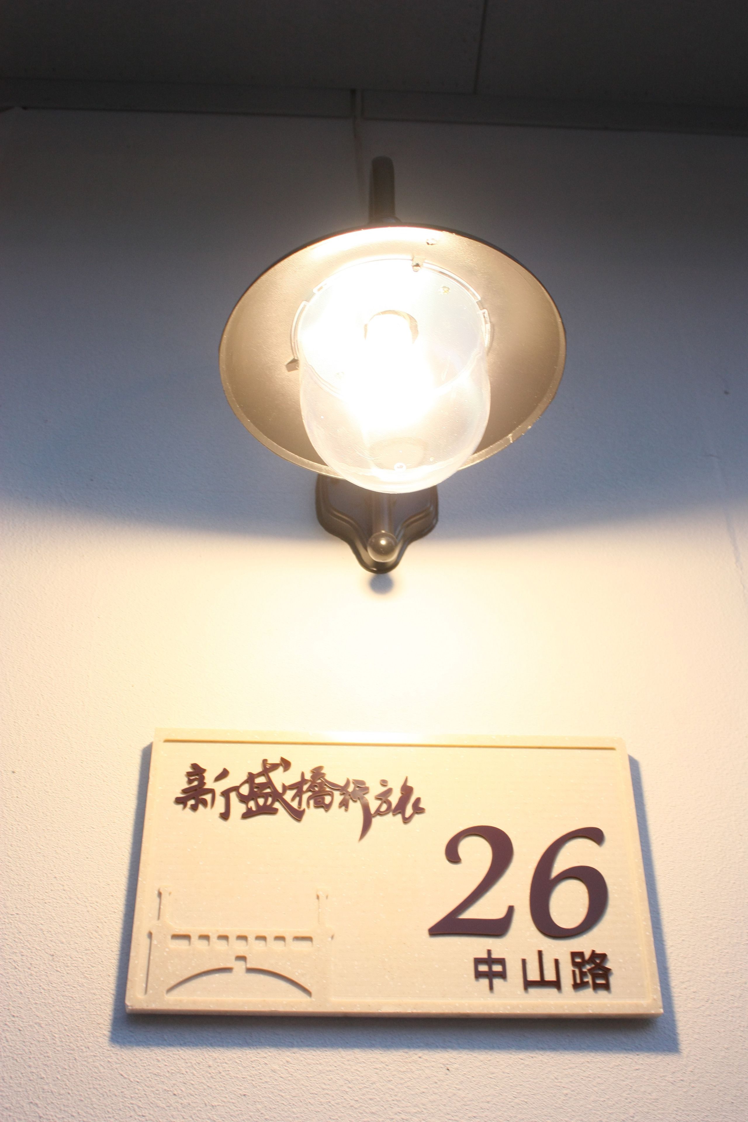 A wall-mounted light fixture with a clear glass cover and a sign below it displaying the number 26 and some text in a foreign script.