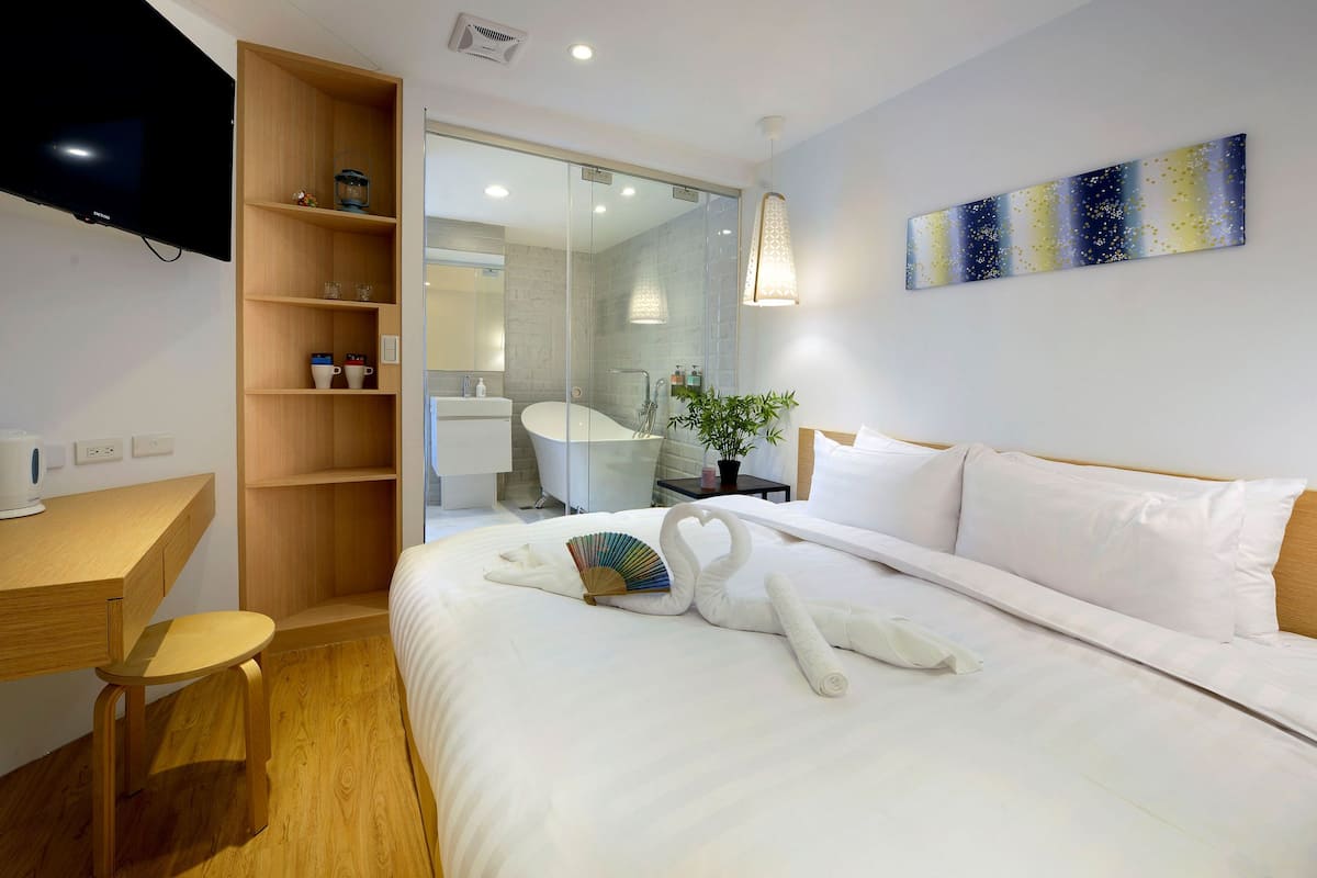A hotel room with a bed, a wooden desk, a television, a glass shower enclosure, and a wall-mounted artwork.