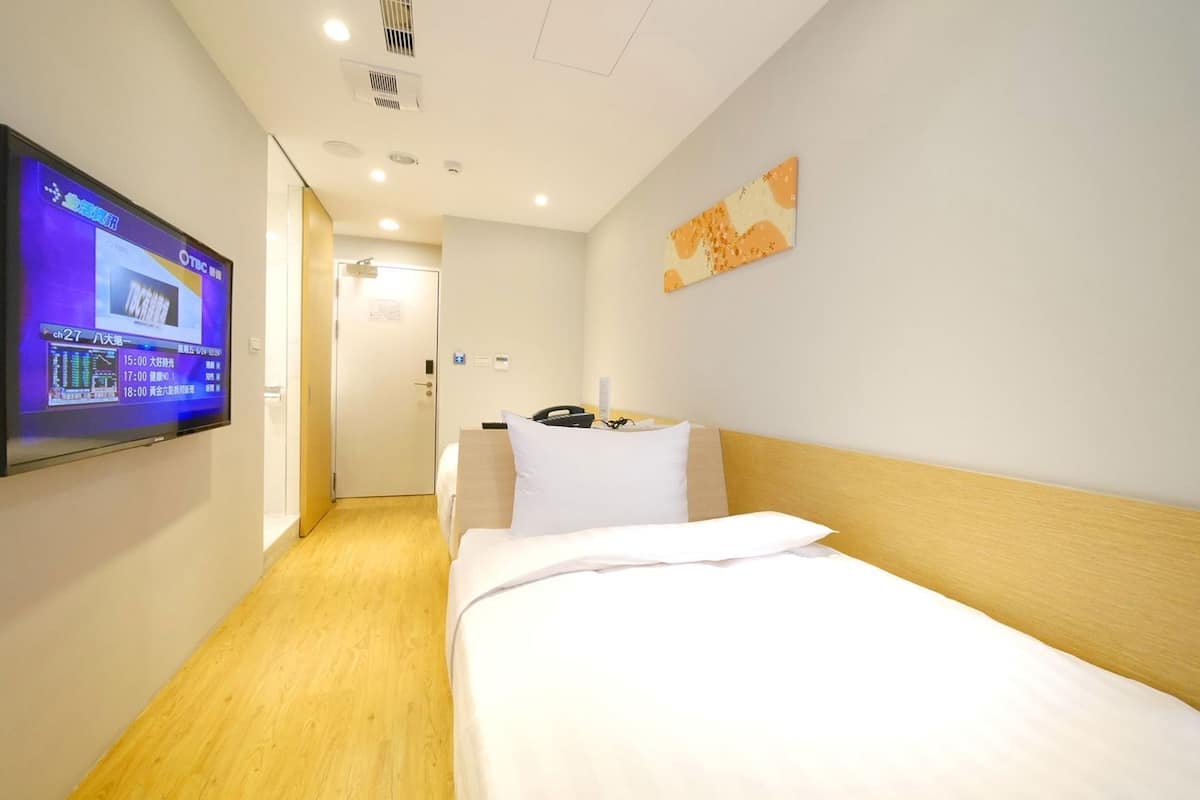 A hotel room with a bed, a TV mounted on the wall, a door, and a small table with a phone and a remote control.