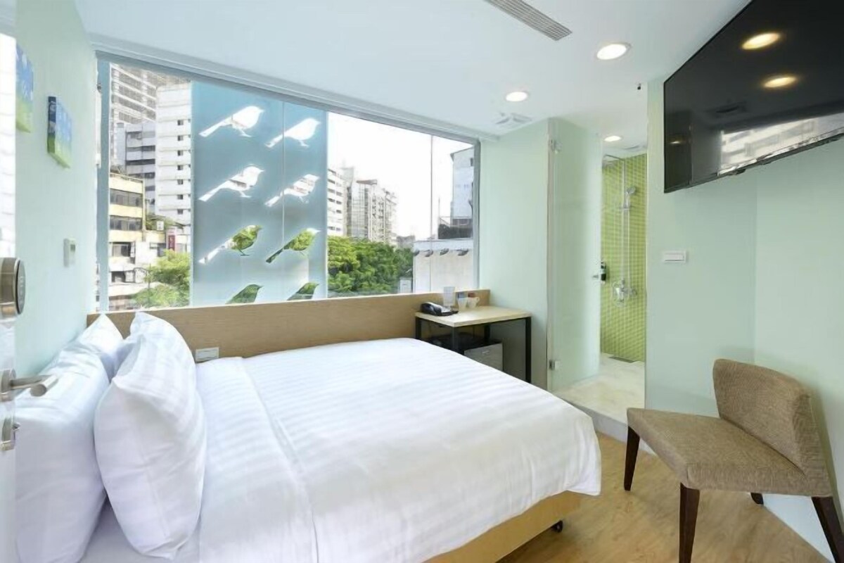 A hotel room with a large bed, a chair, a small table, a TV mounted on the wall, and a view of the city through a large window.