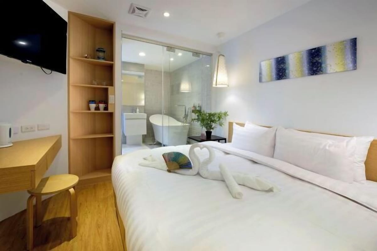 A modern hotel room with a large bed, a wooden desk, a TV, and a bathroom visible through a glass door.