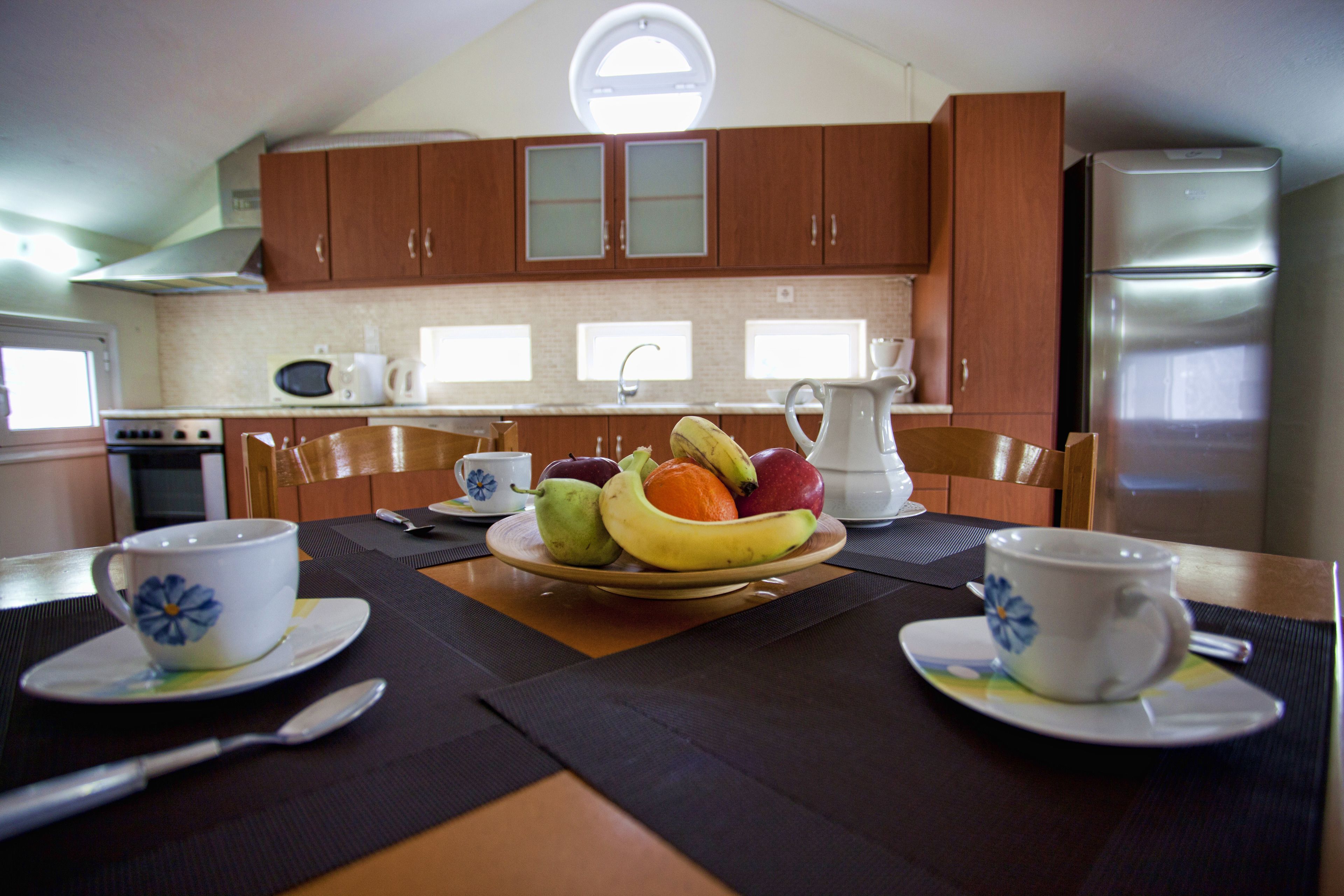 A kitchen with wooden cabinets, a stainless steel refrigerator, and a dining table set with a fruit bowl, two cups, and a pitcher.