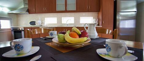 A kitchen with wooden cabinets, a stainless steel refrigerator, and a dining table set with a fruit bowl, two cups, and a pitcher.