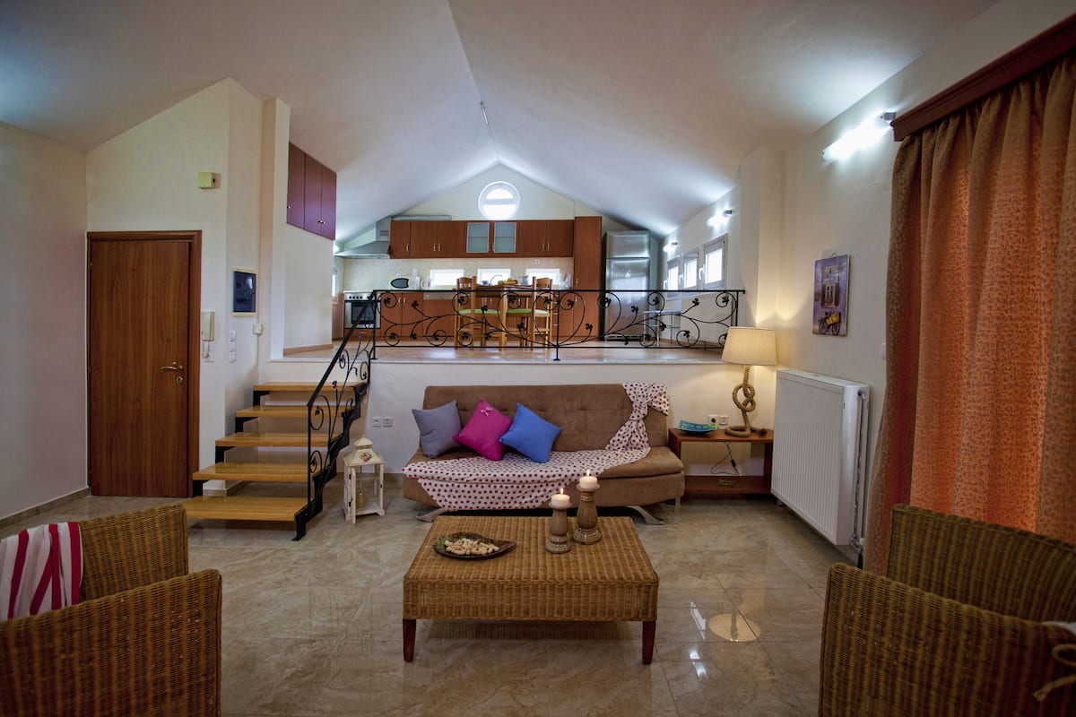 A spacious living area with a high ceiling, a kitchen in the background, and a staircase leading to an upper level.