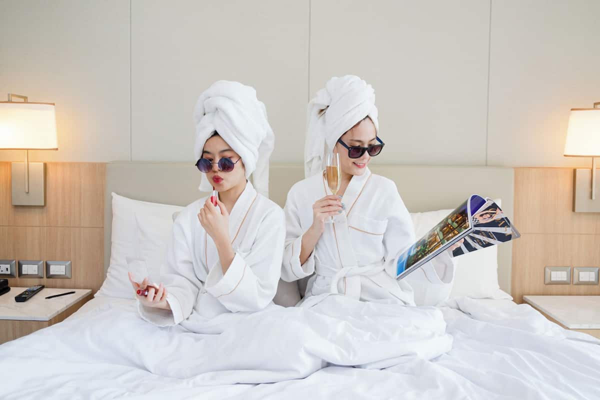 Two individuals in white robes sitting on a bed with a magazine, in a room with a wooden headboard, nightstands, and wall-mounted lamps.