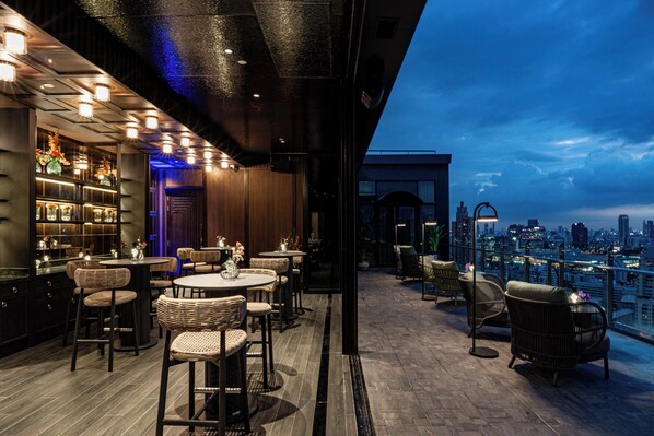 Un bar en la azotea con mobiliario de mimbre, vistas de la ciudad y un interior oscuro con iluminación ambiental.