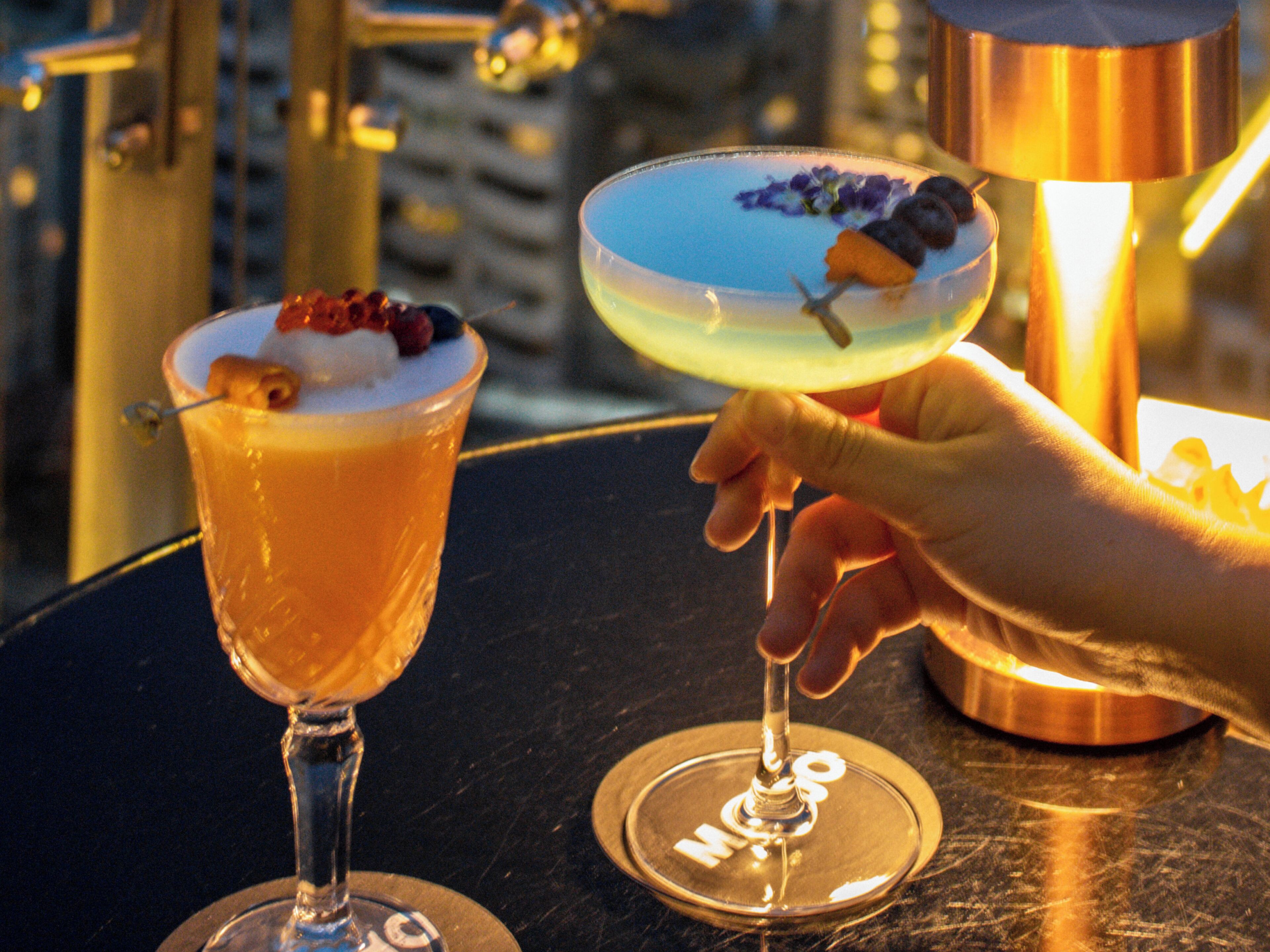 Two cocktails on a table with a cityscape in the background.