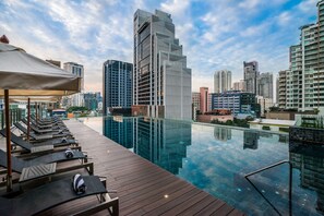 Una piscina infinita en la azotea con sillones de descanso y una terraza de madera, con vistas a un paisaje urbano de edificios modernos.