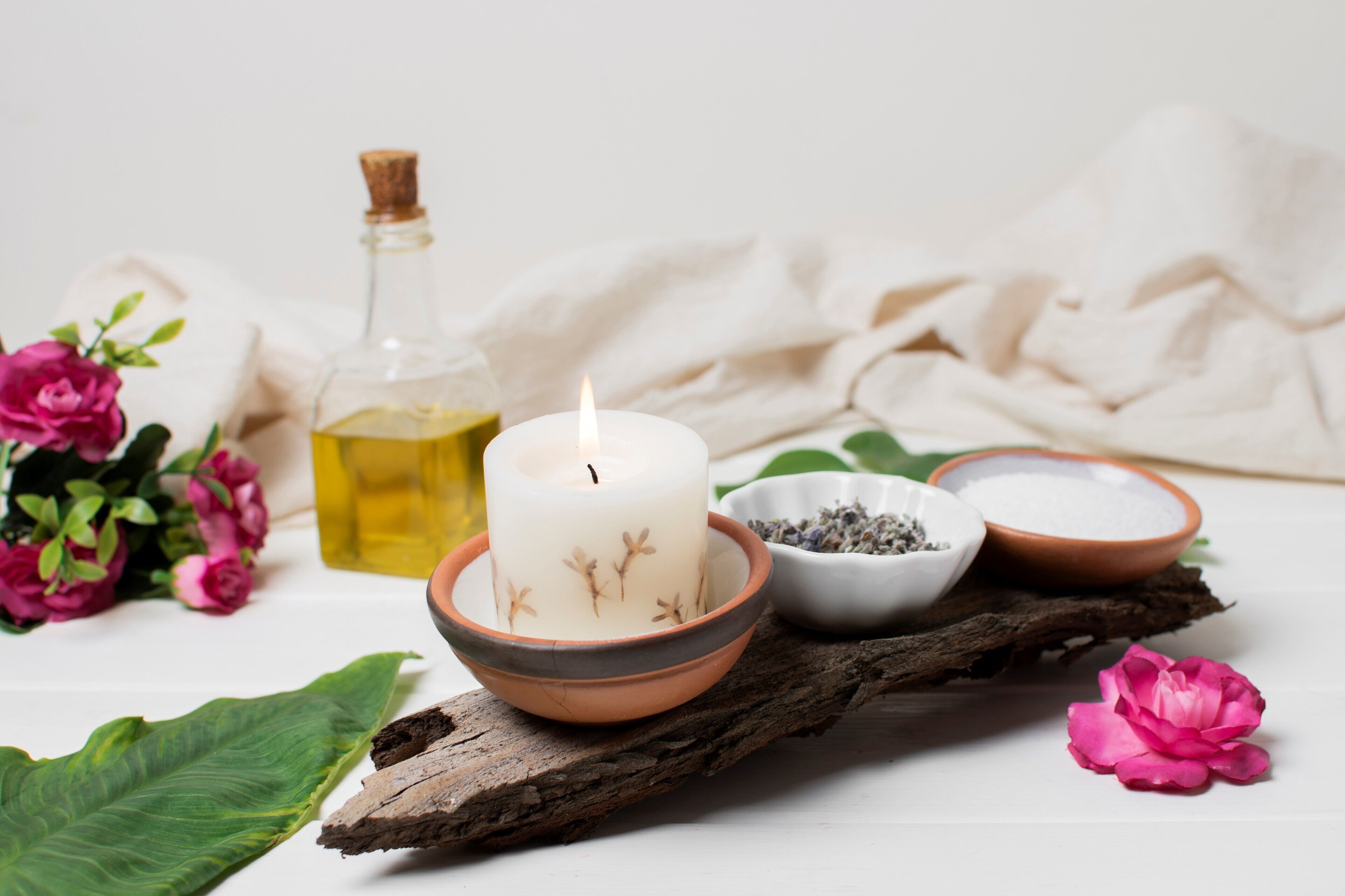 A candle, a bottle of oil, and two bowls with herbs on a wooden surface.