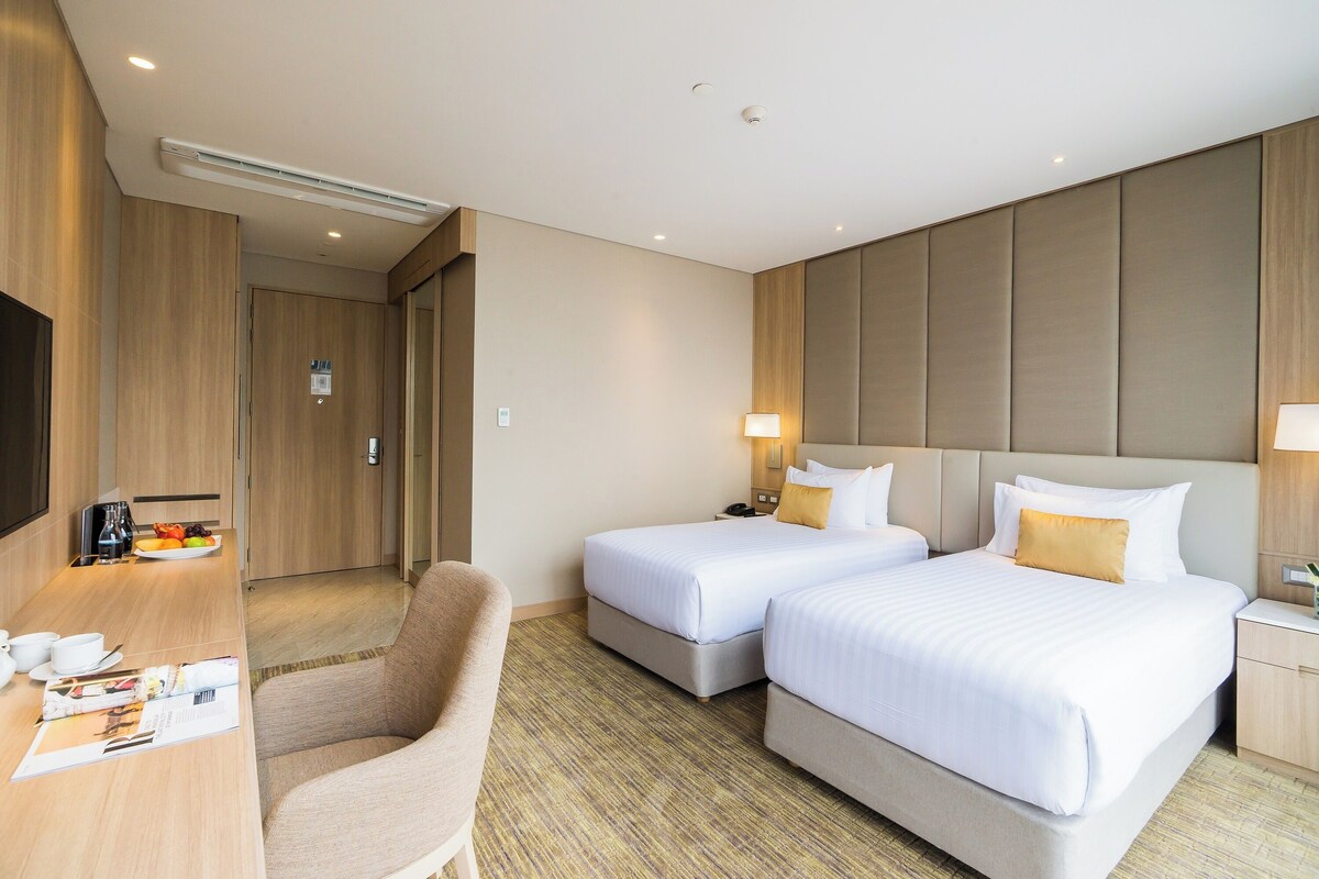 Habitación de hotel con dos camas, un escritorio con silla, televisor y una pequeña zona de comedor con un plato de fruta.