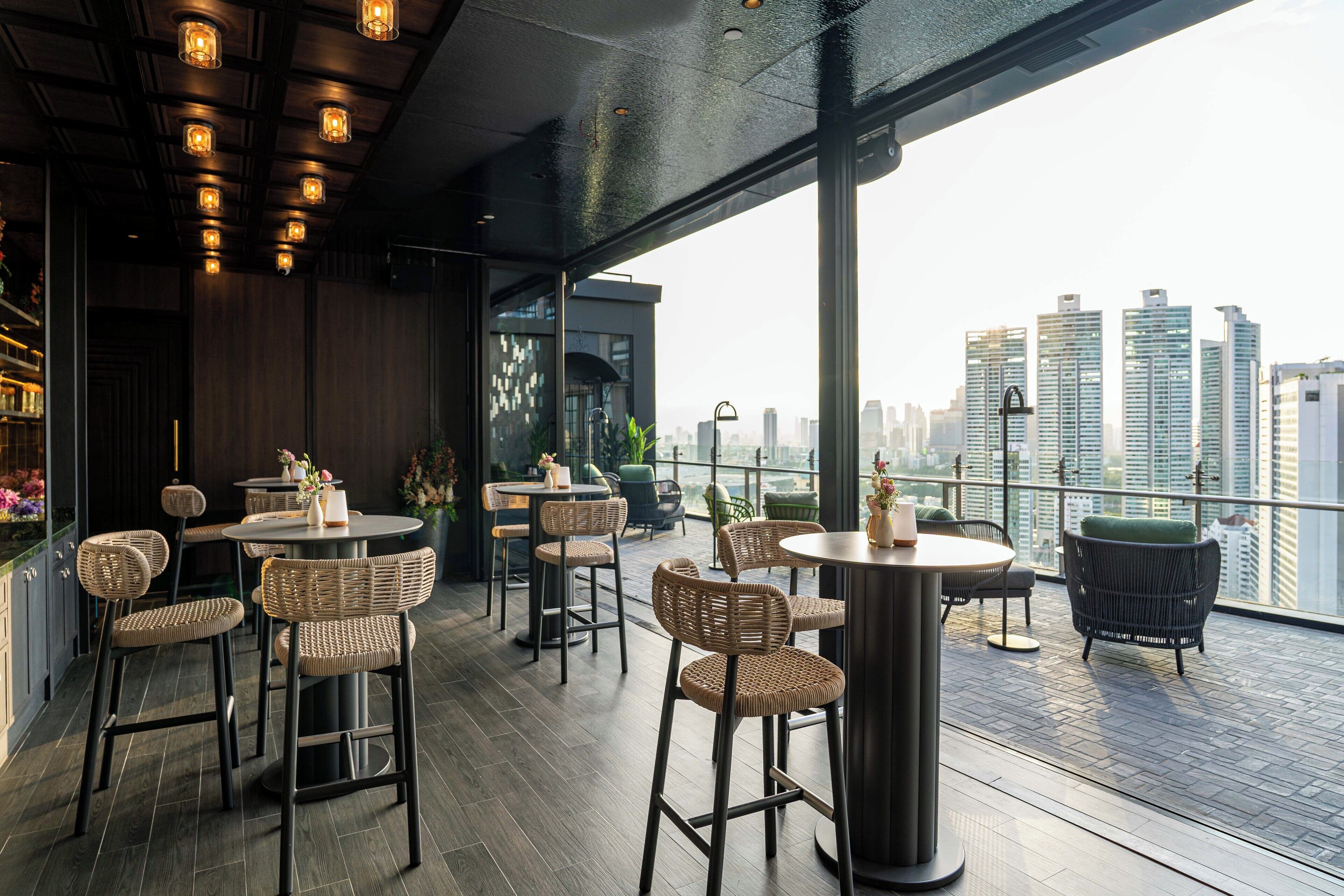 A rooftop bar with wicker chairs and tables, large windows offering a city view, and a modern interior design.