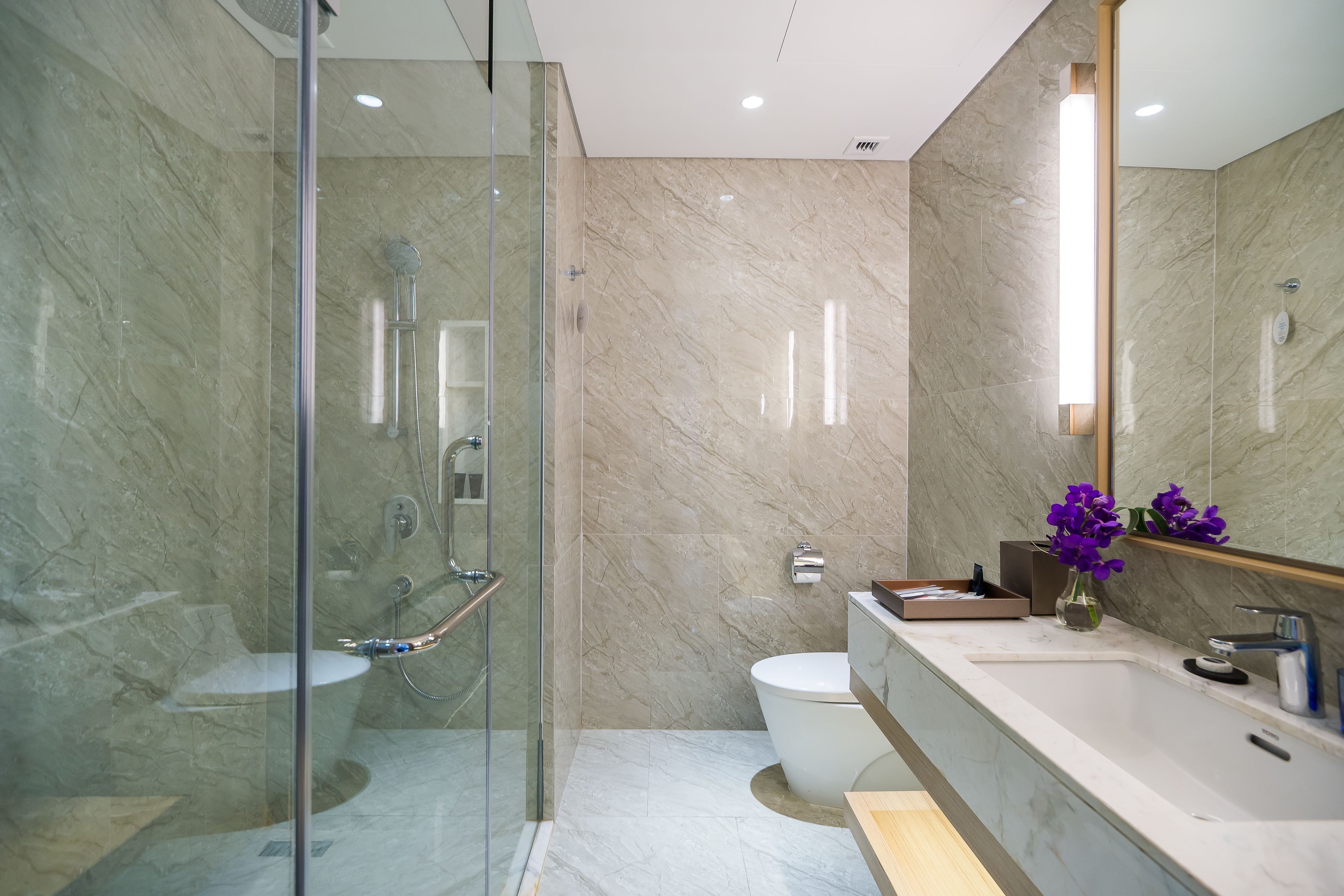 A modern bathroom with a glass-enclosed shower, a marble-tiled wall, a white toilet, and a vanity with a mirror.