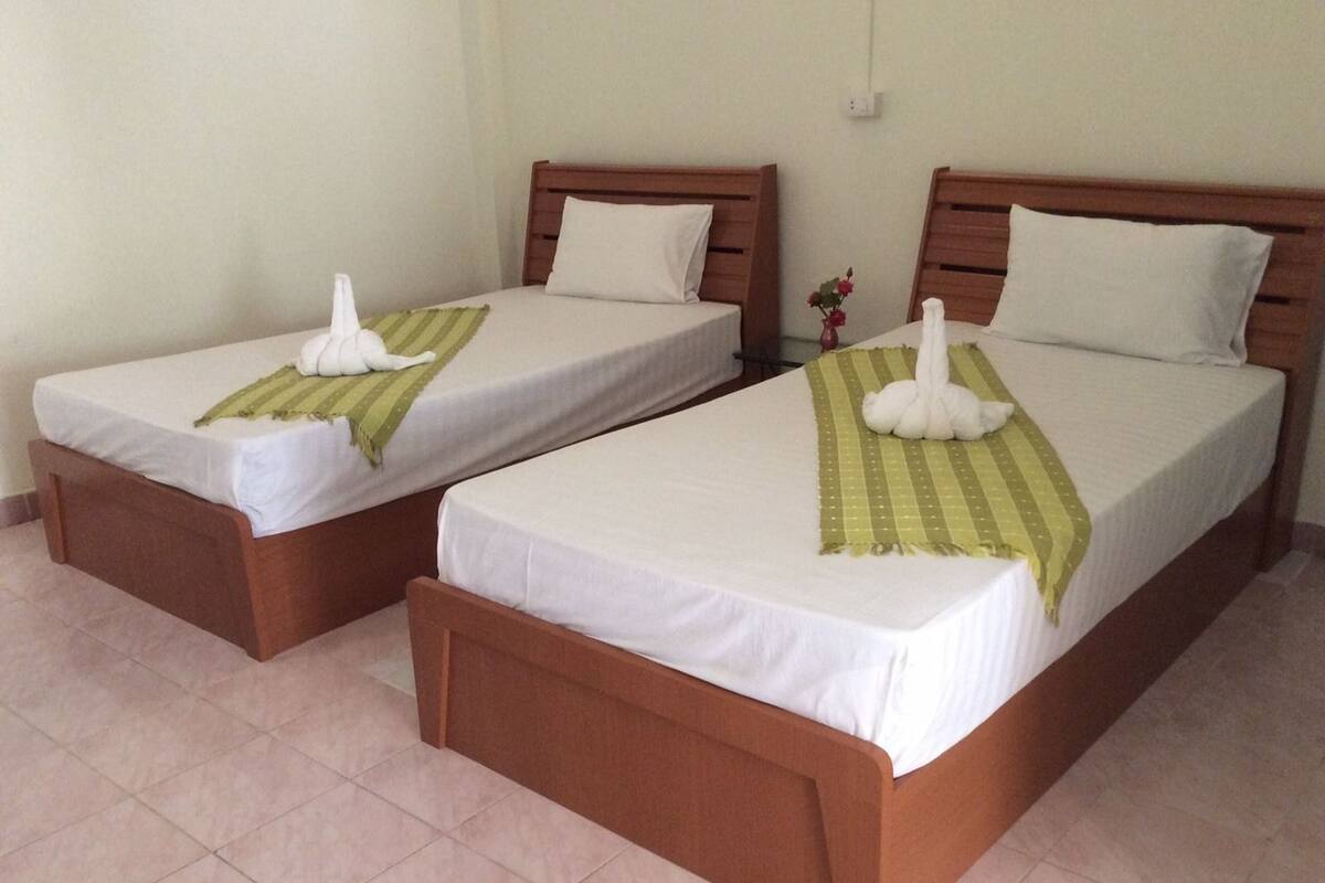 Deux lits simples avec des cadres en bois et du linge de lit blanc, chacun étant équipé d’une serviette et d’un oreiller à rayures vertes.