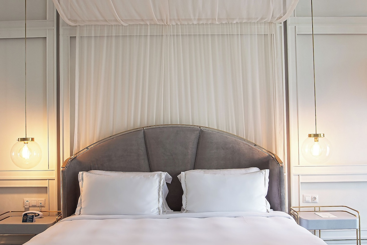 A neatly made bed with white linens, a grey upholstered headboard, and two bedside lamps.
