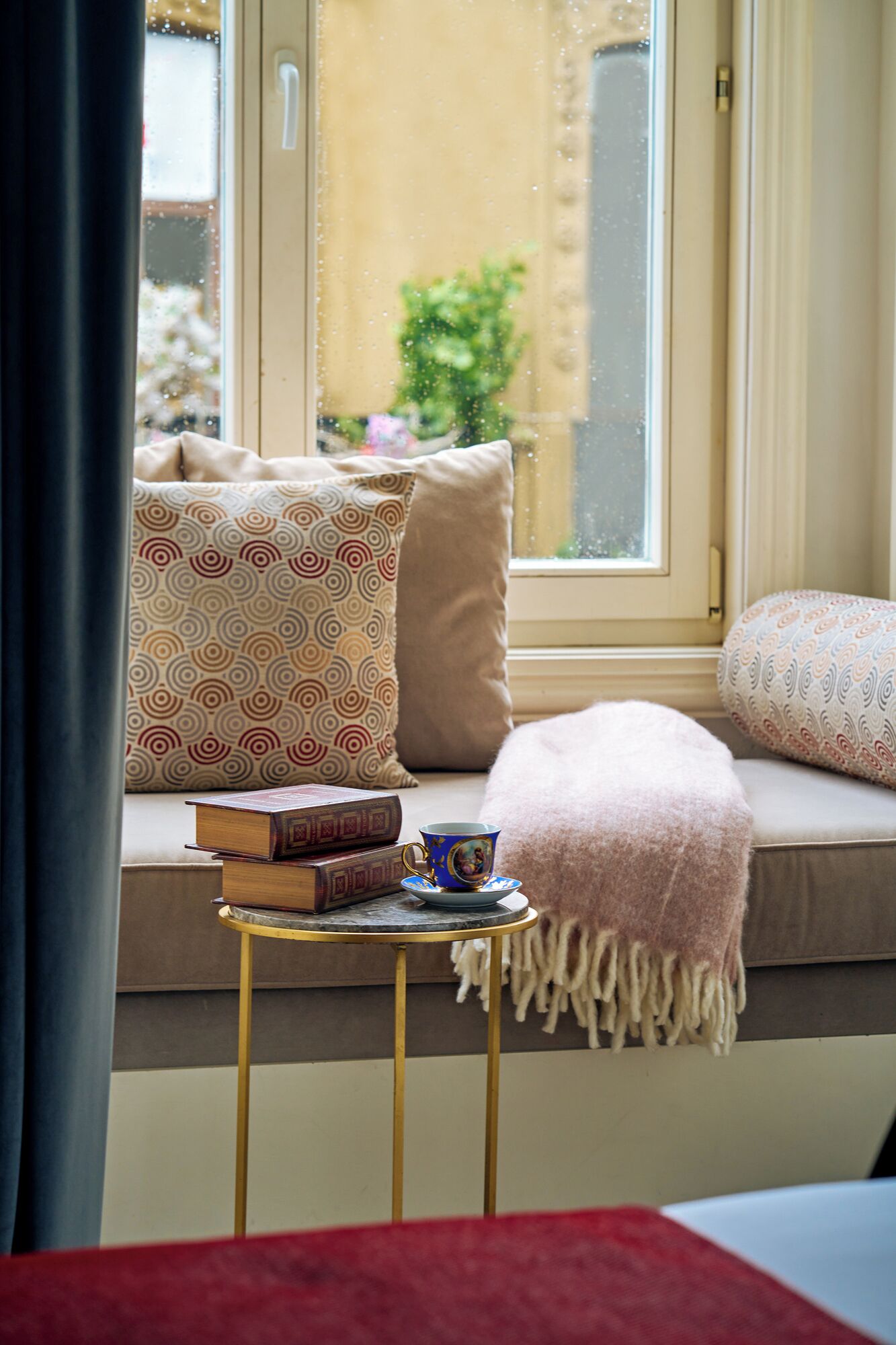 A cozy reading nook with a cushioned bench, a small table with books and a cup, and a view through a window.