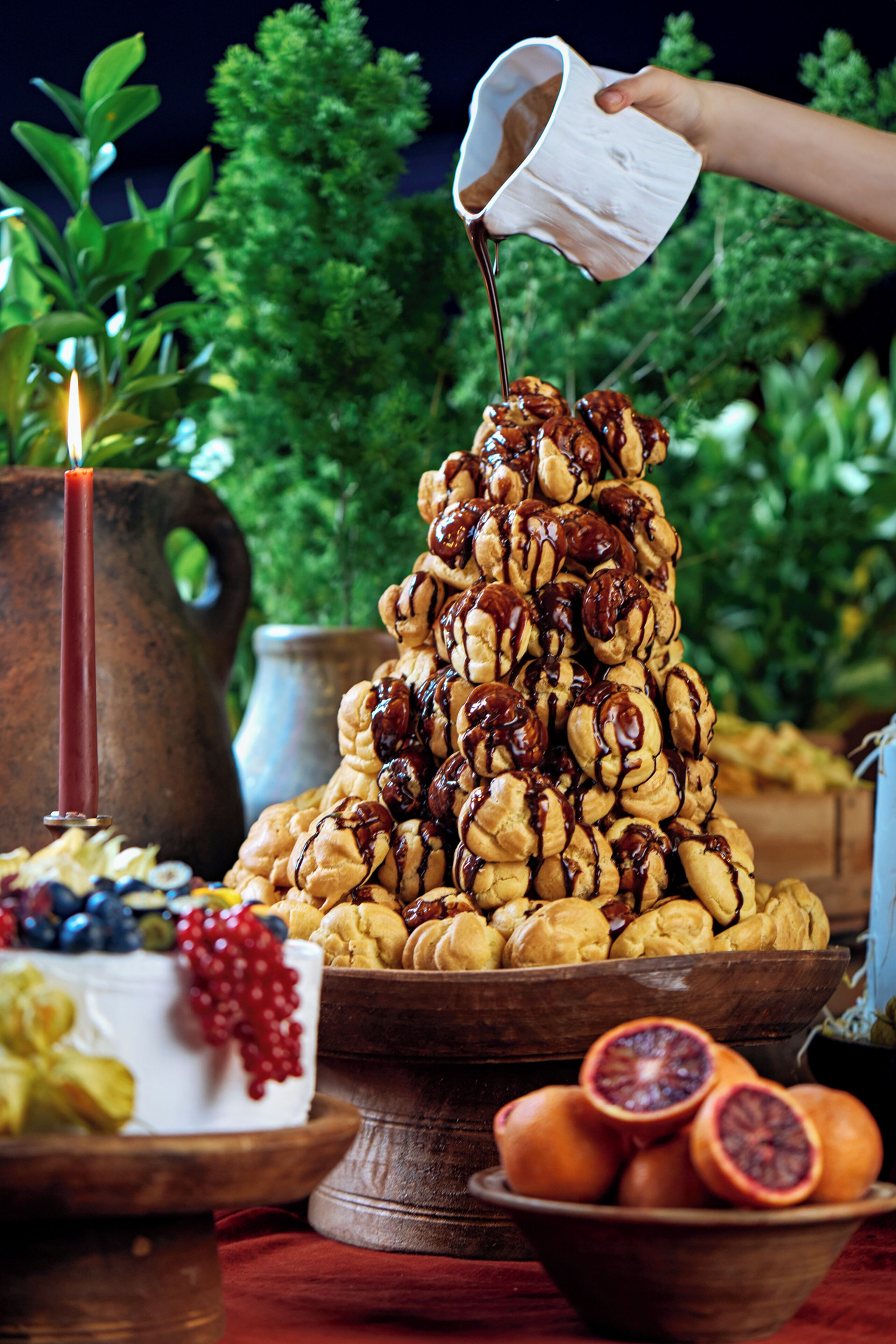 A dessert tower with chocolate sauce being poured over it, surrounded by fruits and a candle.