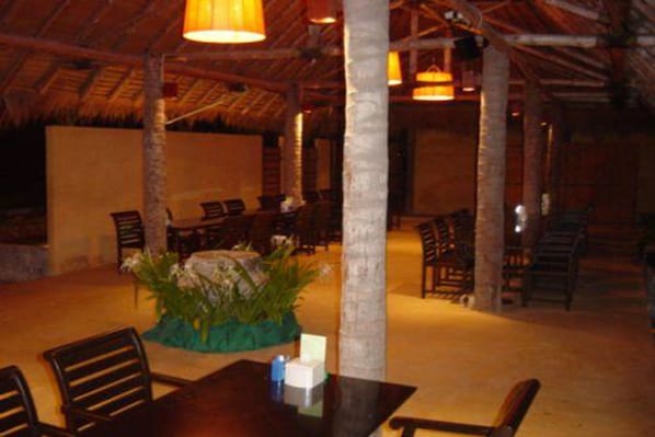 Un comedor interior con mesas y sillas de madera, una planta en el centro y una estructura de techo de paja.