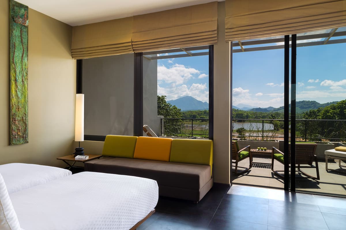 A hotel room with a large bed, a sofa with cushions, a desk with a lamp, and a balcony with a view of mountains and greenery.