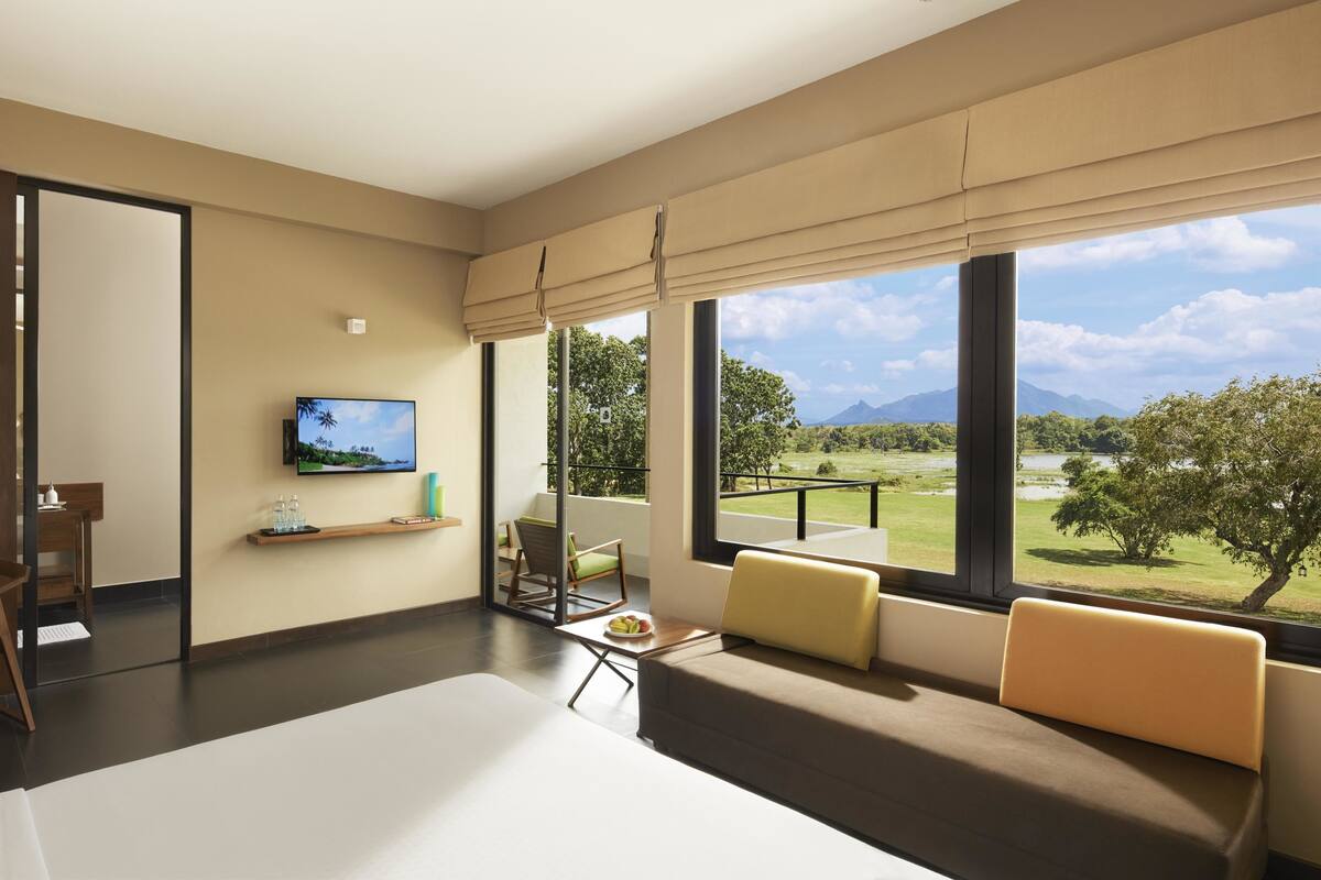 A modern hotel room with a large bed, a TV mounted on the wall, a balcony with a view of the countryside, and a small table with a bowl of fruit.