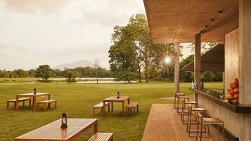 Un espace extérieur aménagé pour les repas, avec des tables et des bancs en bois, un bar avec des tabourets, et une vue sur un lac et des arbres.