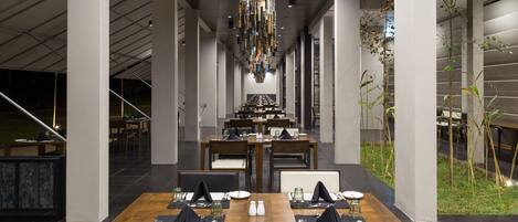 Un comedor moderno con mesas y sillas de madera, un colgante central y vistas al exterior.